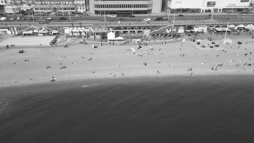 Aerial drone view of busy pebble beach and long promenade with people, seafront buildings and calm water, presented in black and white in Brighton, England, United Kingdom
