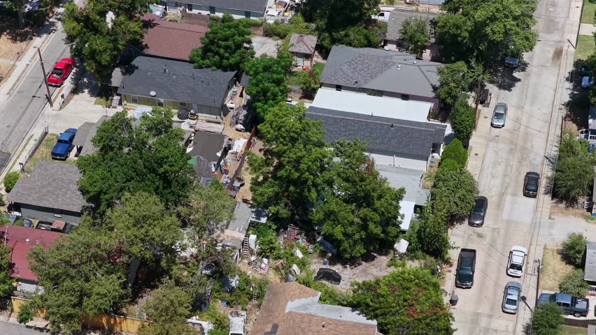 A near-vertical aerial tracks across a West Dallas intersection and adjoining lots. Detached homes, parked cars, driveways, fences, mature trees, utility shadows, and dry patches of grass create a compact geometric study of a residential street crossing.