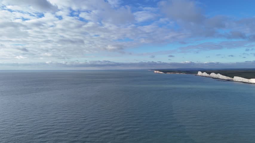 Aerial drone view of Seven Sisters white chalk cliffs extending along the coastline, with calm blue sea and expansive cloudy sky in East Sussex, England, United Kingdom