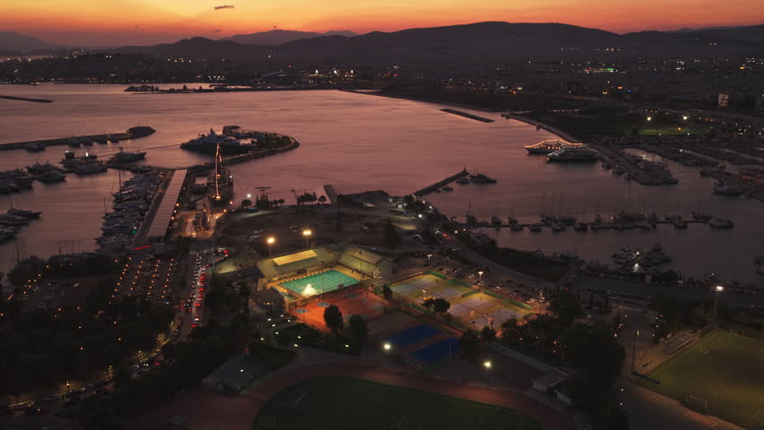 Aerial view tracking over a stunning coastal city of Athens, at sunset, revealing the illuminated marina of Floisvo with luxury yachts, and a modern sports complex featuring swimming pools and courts
