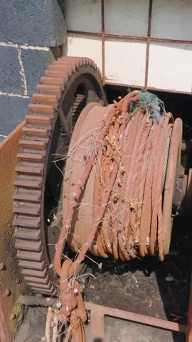 Rusted fishing boat winch with a large cog gear and coiled cable on Hastings beach, England. Camera pans above and then down, showing tangled netting around the rope. Vertical format.