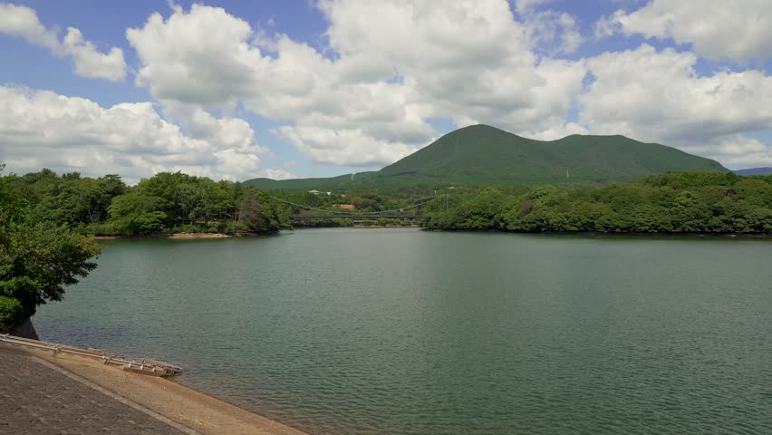 A calm lake stretches across the frame with a suspension bridge spanning the water in the middle distance. Lush green forests line the shores, and a prominent mountain rises under a bright blue sky filled with fluffy white clouds. This peaceful Japanese landscape is ideal for travel, nature, tourism, and lifestyle projects.