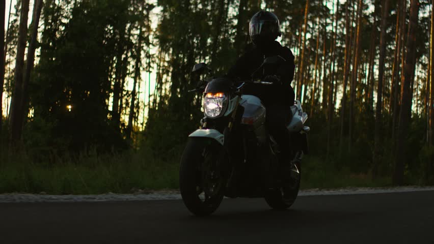 Motorcyclist riding through tranquil forest road at dusk with headlight glowing against tall trees and darkening sky, capturing solitude, speed, and adventure