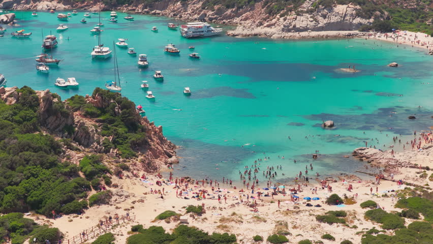 Telephoto aerial of tourists on a beautiful sandy beach nestled in granite cliffs by luxury sailing yachts in turquoise waters of the Isola di Spargi in the Maddalena Archipelago in Sardinia, Italy.