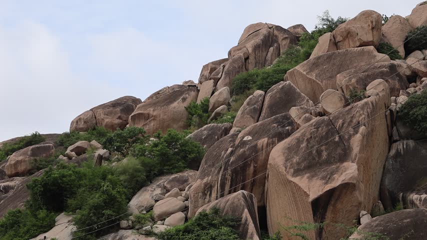 Low-angle shot of a massive rocky hill composed of giant granite boulders stacked naturally, surrounded by green shrubs and trees under an overcast sky