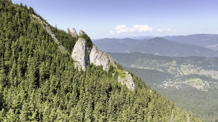Panoramic view of a majestic rocky peak rising from a dense green pine forest in the Ceahlau mountains, with a valley village below.