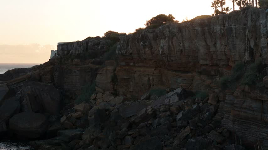 Aerial view of rocky coastal cliffs and a historic fortification at sunset in Cascais, Lisboa, Portugal.