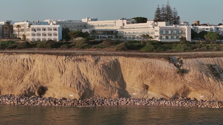 Large white hotel complex sits atop steep sandy cliffs above the calm Atlantic, viewed from a low moving drone offshore.