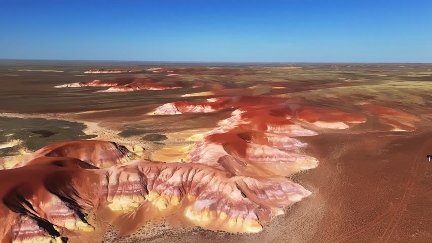 Cinematic drone shot of a solitary figure standing on top of a dramatic colorful red, white, and brown layered rock ridge in Akzhar, Kostanay region, Kazakhstan. The bright summer sunlight illuminates the unique eroded geological strata, contrasting sharply with the surrounding green steppe wilderness