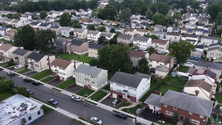 Grid of single-family houses fills dense residential neighborhood in New Jersey, tree-lined streets and parked cars line rows of white and gray rooftops throughout. Aerial drone flyover shot