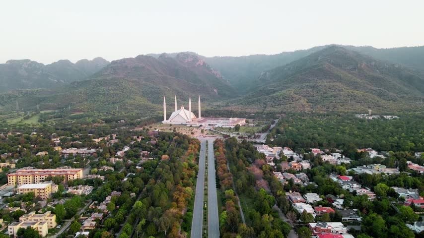 Faisal Mosque stands prominently in this aerial view of Islamabad, Pakistan, showcasing its distinctive modern Islamic architecture, elegant minarets, expansive courtyard, and surrounding Margalla Hills. The elevated perspective highlights this iconic Pakistani landmark.