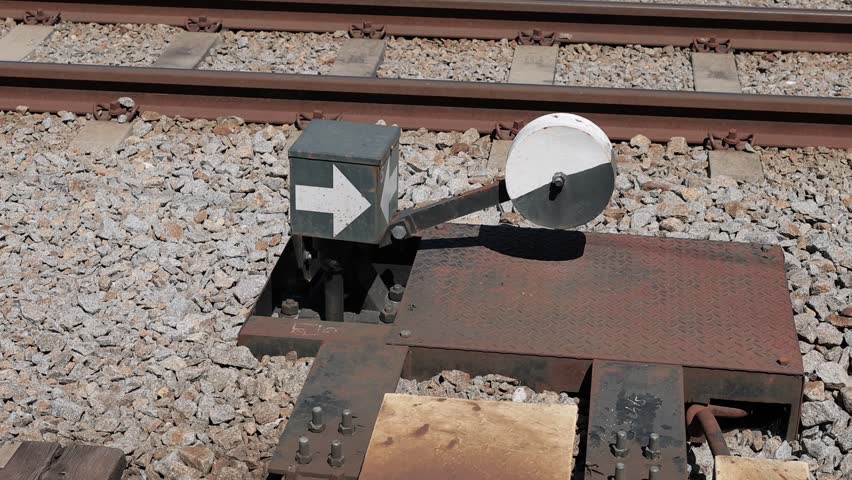 A manual railway switch indicator mechanism featuring a white directional arrow box and a black-and-white counterweight disc, positioned beside train tracks on gravel ballast.