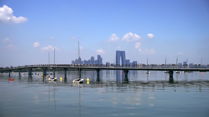 A wide view of a modern city skyline across a calm lake, with a long bridge and small boats on the water under clear blue sky.
