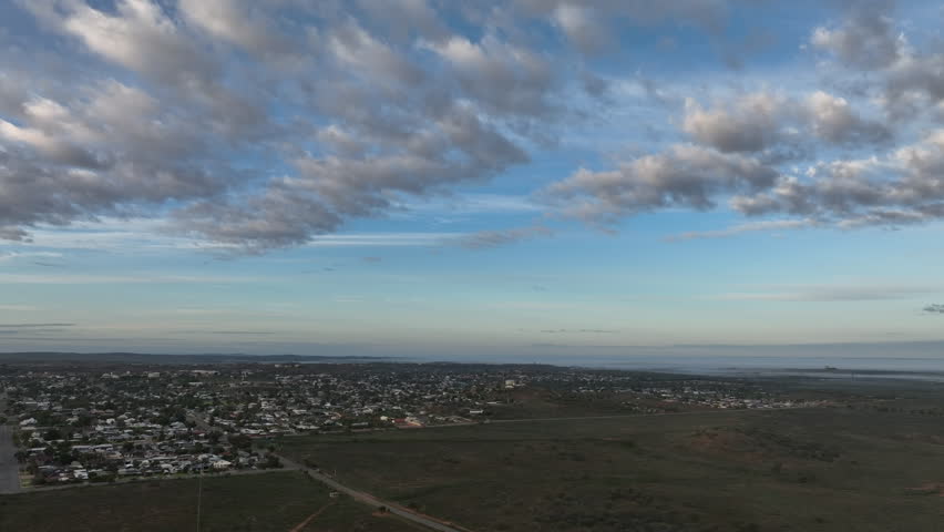 Sweeping aerial establishing view showing the compact urban area of Broken Hill bordered by vast undeveloped terrain beneath a calm blue and cloud-streaked sky.