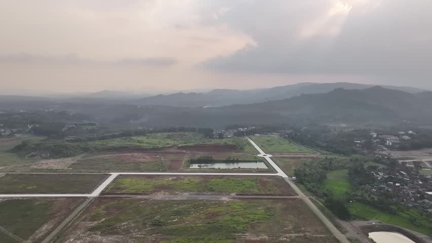 An aerial drone shot captures a vast rural land development project under a cloudy overcast sky, featuring newly graded plots of earth, fresh paved roads, and a small reflective retention pond. Layered mountain ranges fade into the misty horizon in the distance, while patches of green vegetation and exposed red soil create a textured landscape across the expansive valley.