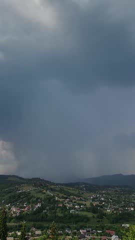 Dramatic weather footage of dark storm clouds gathering above a green mountain village and rolling hills. A heavy rain curtain descends in the distance beneath layered clouds, while scattered houses, winding roads, fields, and forested slopes form a natural rural landscape. Atmospheric footage for weather, climate, nature, travel, documentary, and cinematic projects.