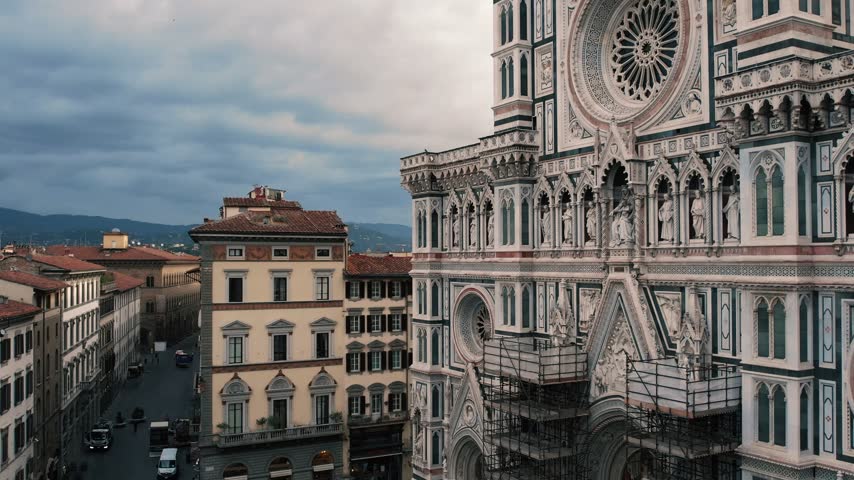 An aerial drone shot ascends past the detailed marble facade of Florence Cathedral, revealing the historic cityscape under a cloudy sky.