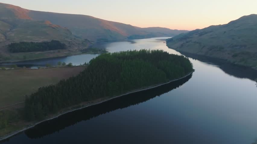 Haweswater Reservoir, English Lake District, Cumbria, UK. Flight towards wooded peninsula of mirror calm lake at sunrise.