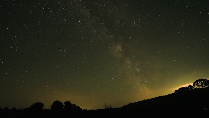 Wide Milky Way Timelapse of Galactic Center as near Earth Satellites Pass By Overhead with Silhouetted Earth Landscape Below.