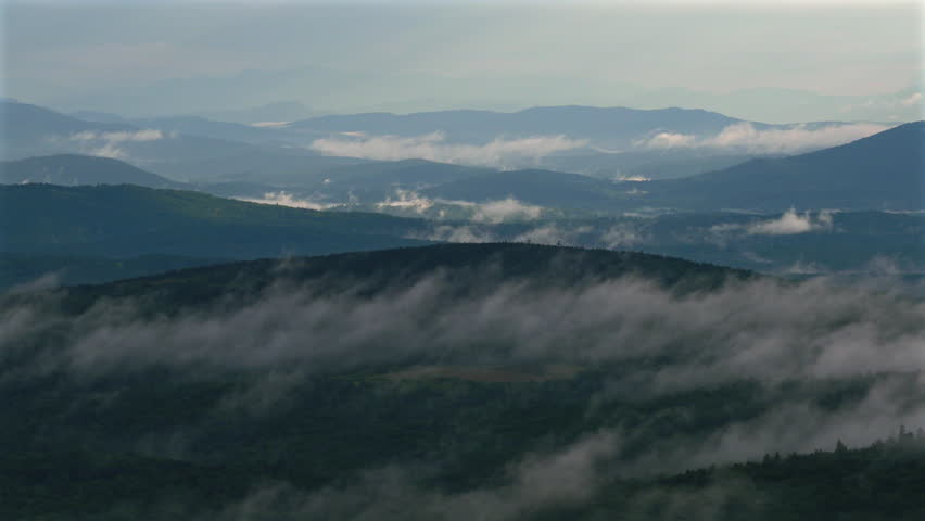 Sideways aerial movement reveals fog drifting through forested valleys and layered mountain ridges across rural New Hampshire lakes region, with a compressed telephoto perspective towards the sunlight