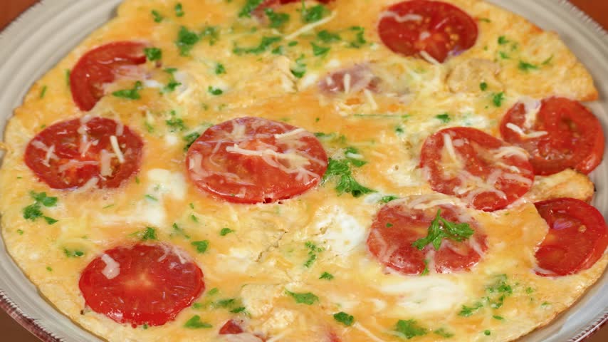 A cooked omelet is topped with sliced tomato, parsley and melted cheese. The rotating presentation highlights tomato and parsley plants in a colorful protein meal.