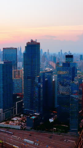 Dusk skyline of Long Island City towers, Queens, New York. Elevated drone perspective of glass high rises, train yard and pink sunset clouds. Vertical video.