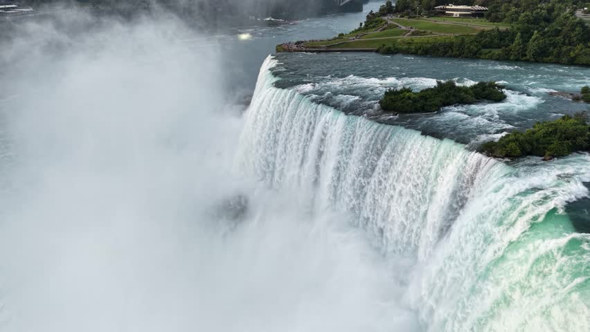 Wide aerial tracking shot moving forward over a thundering vertical water plunge, green rocky islands, and massive plumes of white mist at the crest of the Horseshoe Falls, Canada