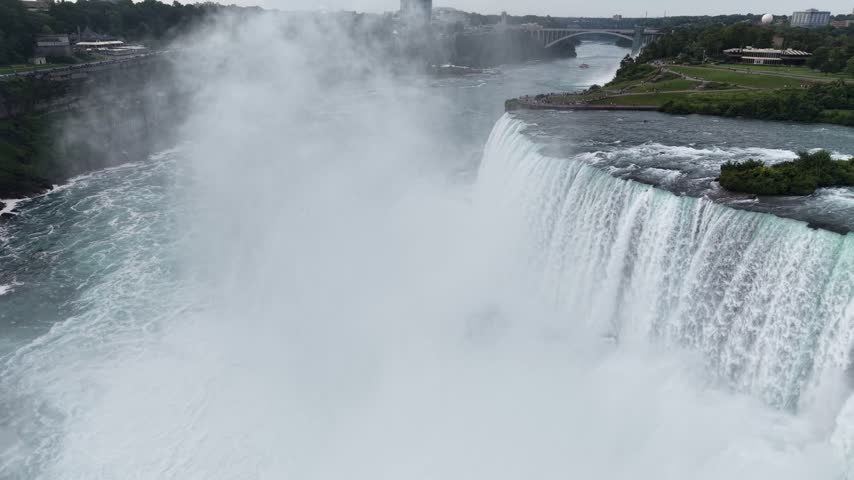 An aerial drone shot flies forward through heavy rising mist over the rushing crest of Horseshoe Falls, revealing the distant Rainbow Bridge stretching across the Niagara River gorge
