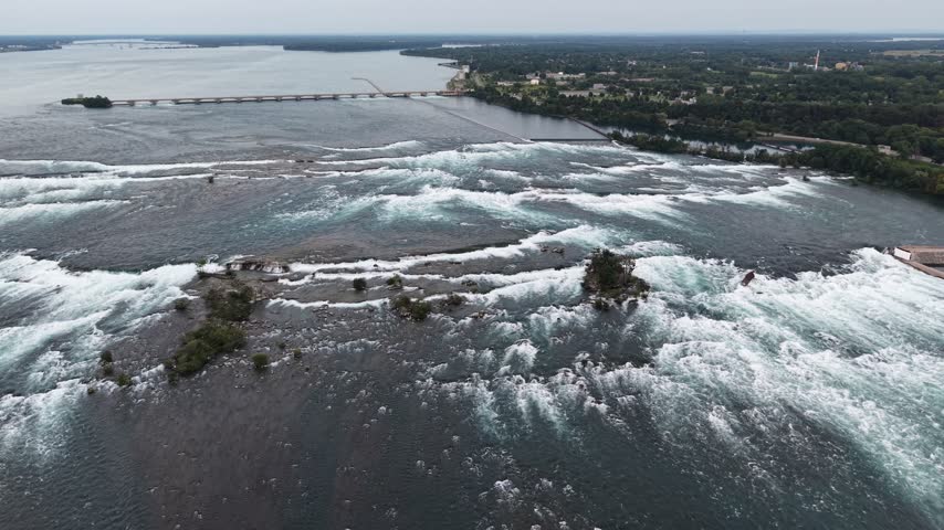 An aerial drone shot flies forward over the churning white water rapids of the upper Niagara River, revealing the International Control Dam and the distant lake horizon
