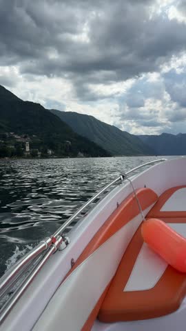 Scenic view from an orange and white motorboat cruising across Lake Como, surrounded by green mountains beneath a dramatic cloudy sky in Italy.