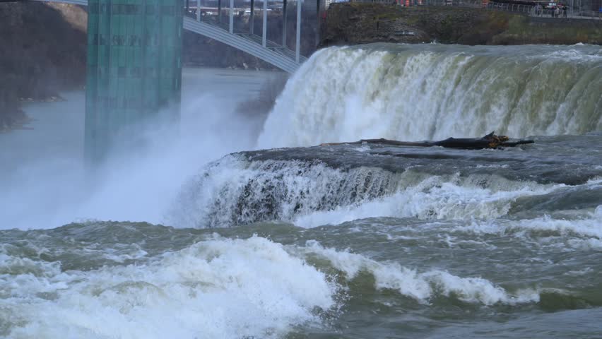 Panoramic view of Niagara Falls and the turbulent rapids of the Niagara River. A powerful flow of water moves rapidly toward the waterfall, creating dense water mist. In the background, the Rainbow Bridge, pedestrian infrastructure, and an observation tower are visible within a scenic landscape.