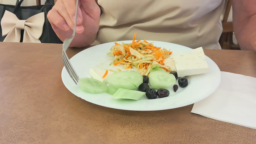 A person is eating a healthy plate of sliced cheese, fresh cucumbers, olives, and a carrot salad.