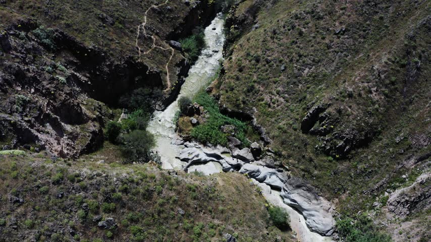 Aerial drone view of a mountain river flowing through a narrow rocky canyon surrounded by steep rugged slopes, exposed stone formations and patches of green vegetation. The water follows a winding natural channel through eroded rock, creating a dramatic remote landscape shaped by water and geological erosion