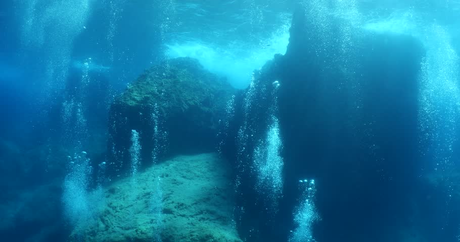  scuba divers exploring around a reef underwater deep blue water big rocks and bubbles ocean scenery 