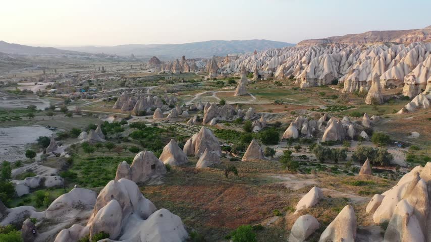 Panoramic aerial drone view of white rock formations, conical fairy chimneys, and flat-topped tuff mountains in Rose Valley, Cappadocia, Nevsehir, Turkey under a clear blue sky.