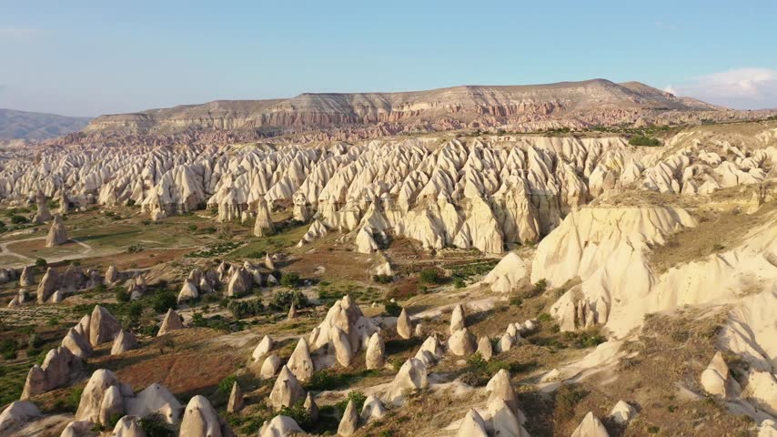 Panoramic aerial drone view of white rock formations, conical fairy chimneys, and flat-topped tuff mountains in Rose Valley, Cappadocia, Nevsehir, Turkey under a clear blue sky.