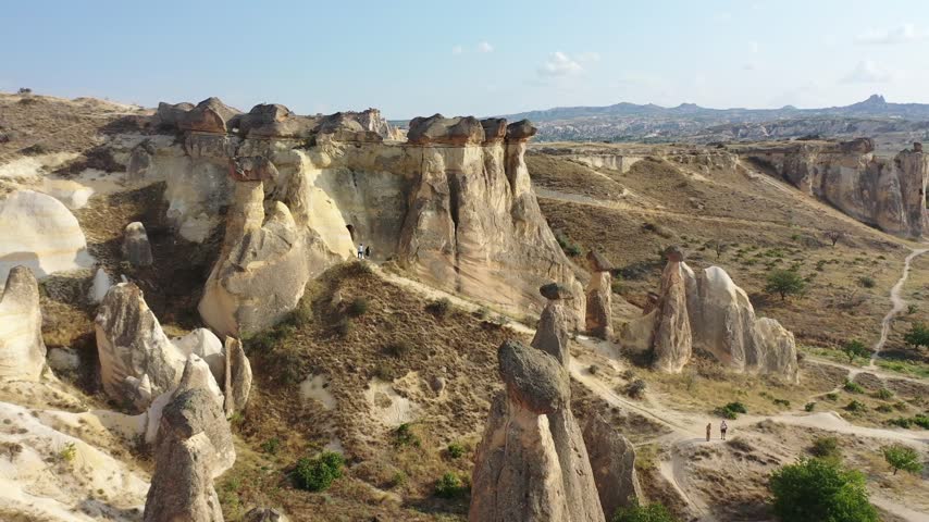 Panoramic aerial drone view of white rock formations, conical fairy chimneys, and flat-topped tuff mountains in Rose Valley, Cappadocia, Nevsehir, Turkey under a clear blue sky.