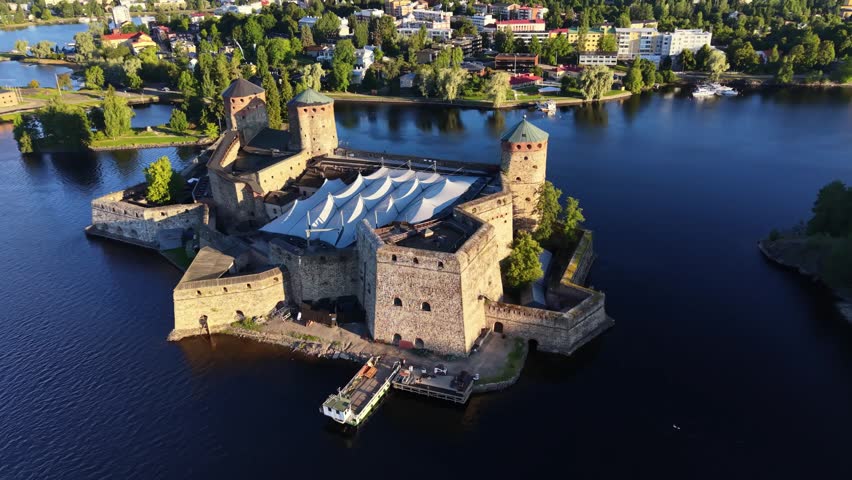 Aerial panoramic view of medieval Olavinlinna Castle surrounded by Lake Saimaa under a blue sky in Savonlinna, Finland.