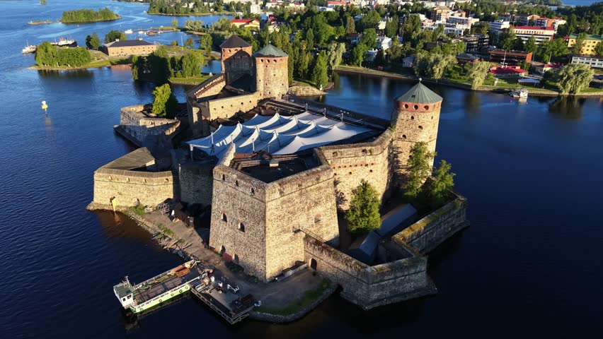 Aerial panoramic view of medieval Olavinlinna Castle surrounded by Lake Saimaa under a blue sky in Savonlinna, Finland.