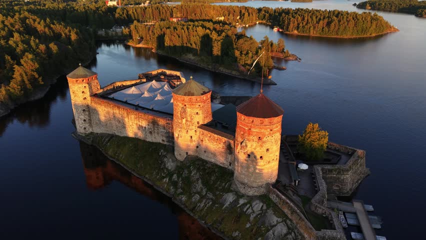 High aerial view of medieval Olavinlinna Castle illuminated by golden hour sunset light on Lake Saimaa in Savonlinna, Finland.