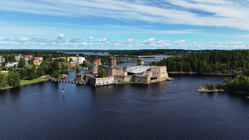 Aerial panoramic view of medieval Olavinlinna Castle surrounded by Lake Saimaa under a blue sky in Savonlinna, Finland.