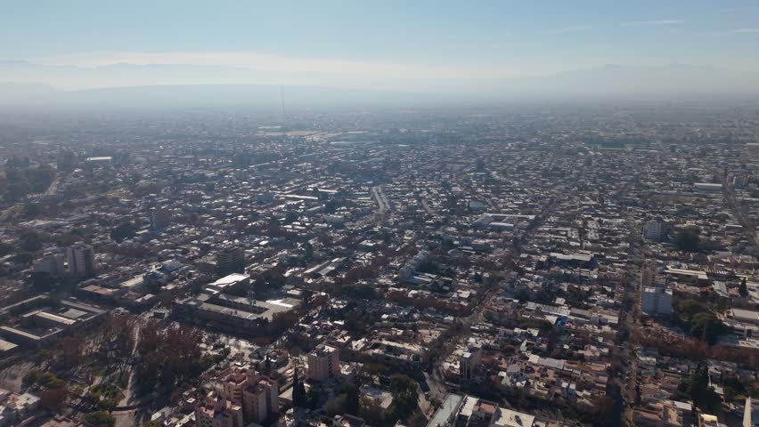 Aerial view of San Juan Capital, Argentina, showcasing dense urban sprawl, grid streets, and distant Andes mountains under crisp daylight with gentle haze.