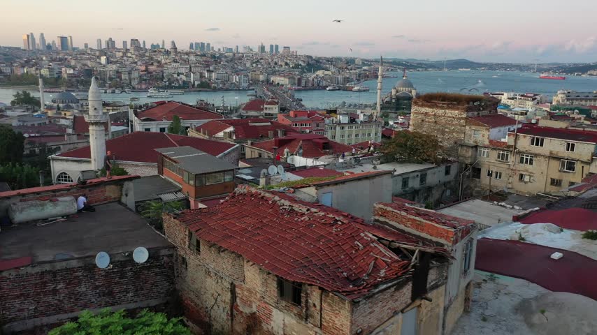 High aerial drone shot over old Istanbul red tile rooftops overlooking mosques, the Golden Horn waterway, Galata Bridge, and the modern city skyline during dusk twilight in Turkey.