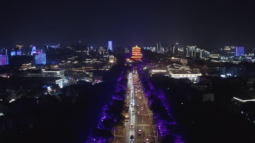 High aerial drone view of the brightly illuminated iconic Yellow Crane Tower glowing golden red at night surrounded by modern city buildings and traffic in Wuhan, Hubei province, China.