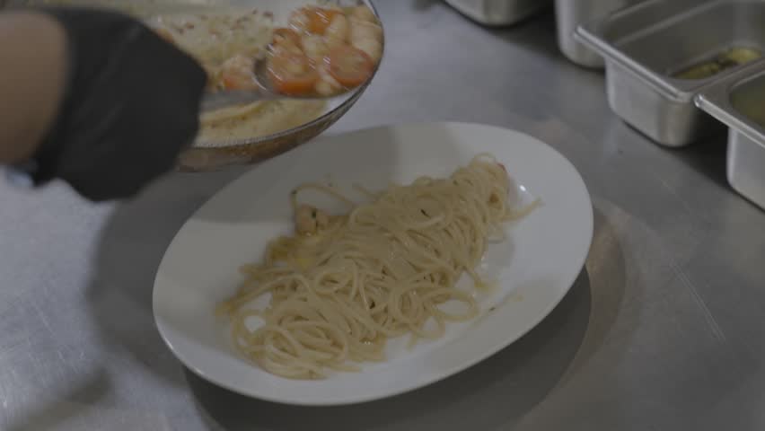 Chef plating freshly cooked spaghetti with shrimp and cherry tomatoes in a restaurant kitchen, showing professional pasta preparation and food service.