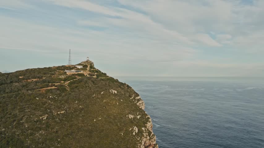 Drone orbit shot circles rugged Cape of Good Hope coastline in South Africa, revealing green cliffs, ocean waves, lighthouse, and expansive sky in scenic coastal landscape with travel appeal.