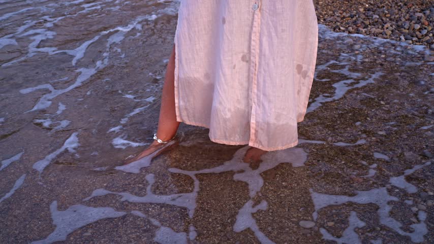 A woman stands in shallow water at a beach. She enjoys the feeling of waves touching her feet. The sun sets in the background, creating a warm glow on the sand.