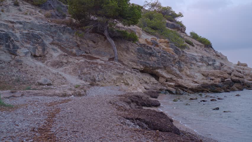 Footsteps on a rocky beach near calm water. The sky changes colors as the sun sets over the distant hills. Waves gently hit the shore and nearby rocks.