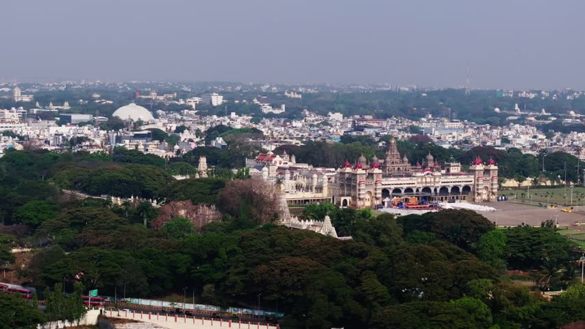 Wide aerial shot showcasing ornate architecture of Mysore palace. Cinematic footage flying over lush green trees surrounding historic landmark in Mysore city landscape with clear sky.