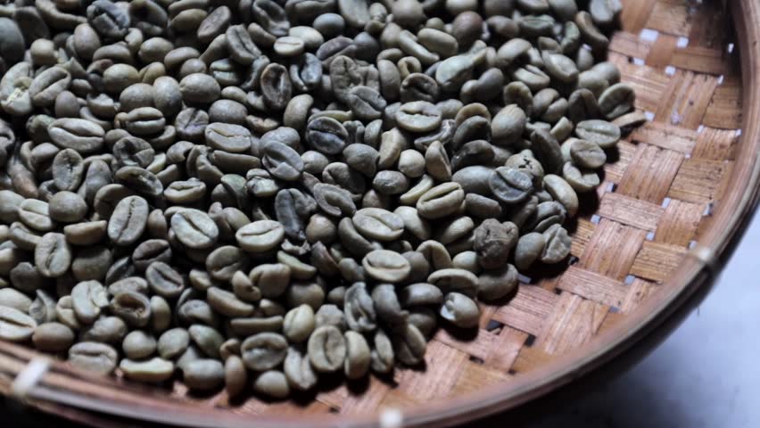 High angle slow motion video of raw green coffee beans piled inside a round traditional woven bamboo basket. Organic unroasted green coffee beans arranged in a rustic wooden wicker tray captured in slow motion. Slow motion detailed shot of natural raw arabica coffee beans placed on a textured woven background.